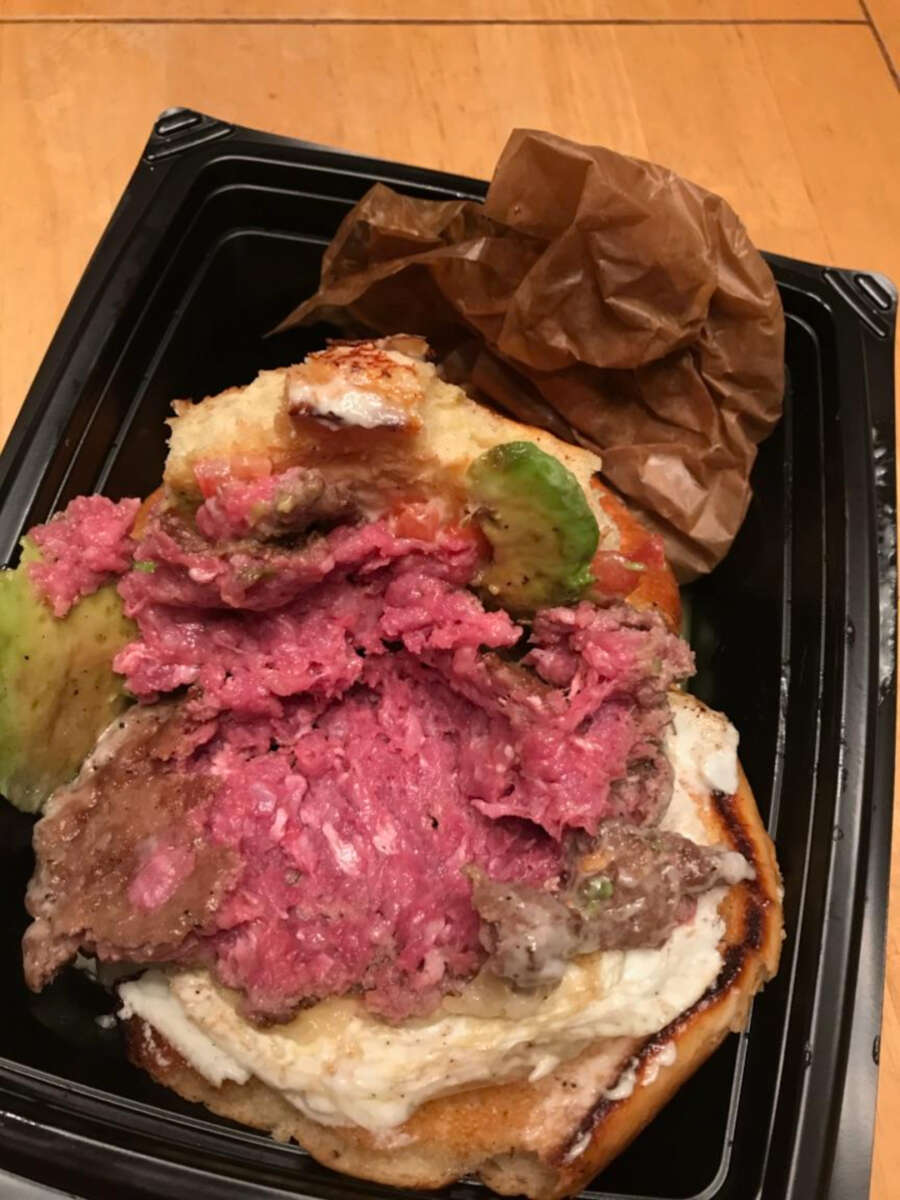 A burger with a bun, sliced avocado, and a patty that appears very undercooked and pink inside, displayed in a black takeout container on a wooden table.