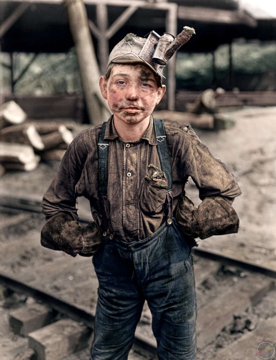 A young boy coal miner stands on a dirt ground by railway tracks, wearing dirty work clothes, suspenders, gloves, and a headlamp helmet, with a tired, soot-stained face and hands on his hips.