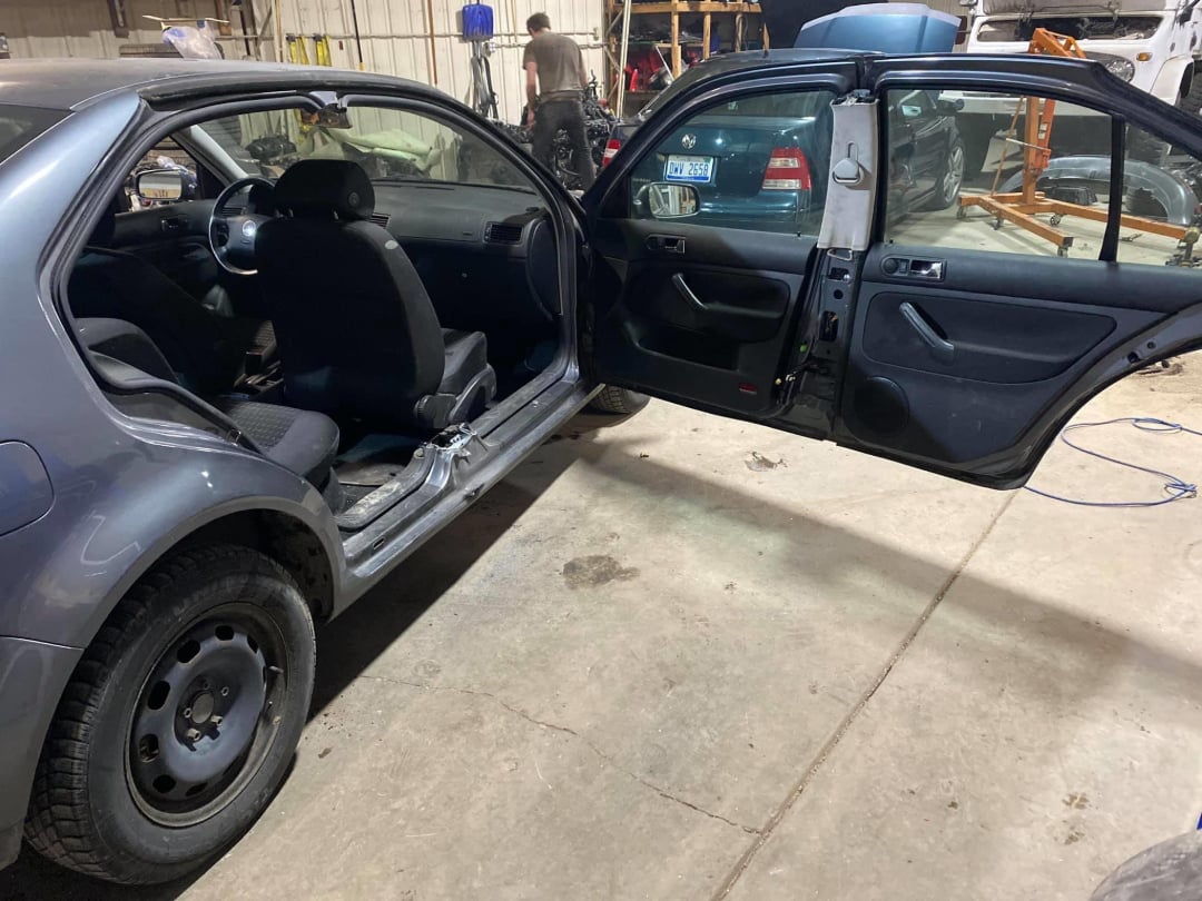 A car in a garage with its rear door and front passenger door open, showing missing parts like the rear seat and interior trim. Another car and workshop tools are visible in the background.