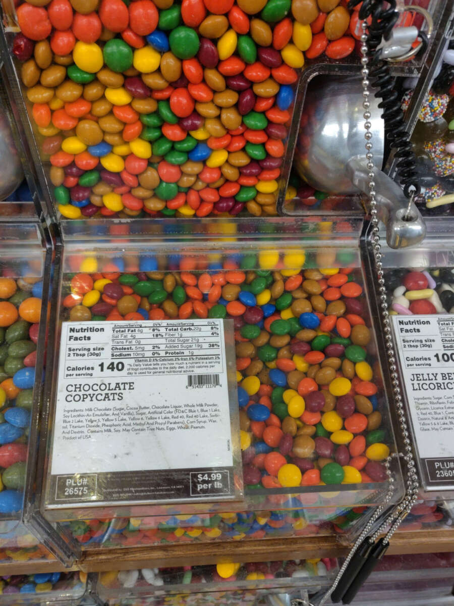 A bulk bin filled with colorful candy-coated chocolate pieces labeled “Chocolate Copycats,” with a scoop attached and a nutrition facts label visible on the front.