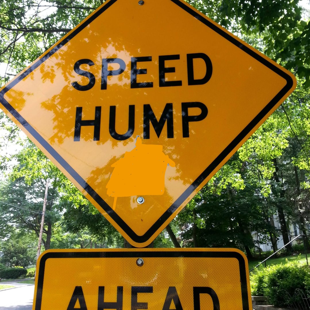 A yellow diamond-shaped road sign reads "SPEED HUMP" with a section in the middle obscured by orange paint. Green trees and a street are visible in the background.