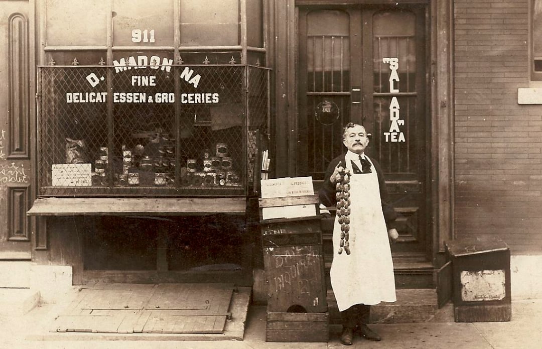 A man wearing a white apron stands in front of a deli storefront, holding a string of garlic. Signs in the window read “Fine Delicatessen Groceries” and “Tea.” The shop window displays various goods.