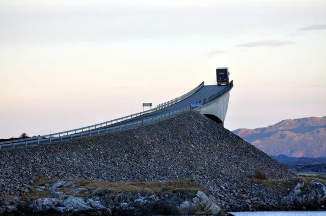 A truck drives over a sharply curved bridge that rises steeply above rocky terrain, with mountains and a pastel sky in the background.
