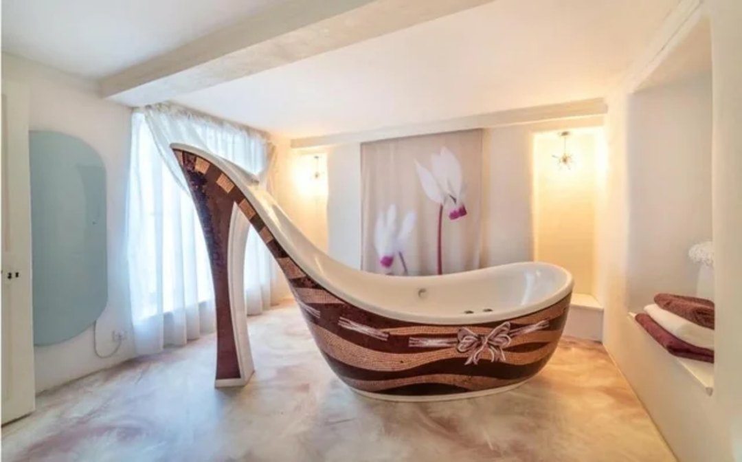 A unique bathroom features a bathtub shaped like a high-heeled shoe, with brown and beige mosaic patterns. The room is softly lit, with white curtains, folded towels, and a floral wall art in the background.