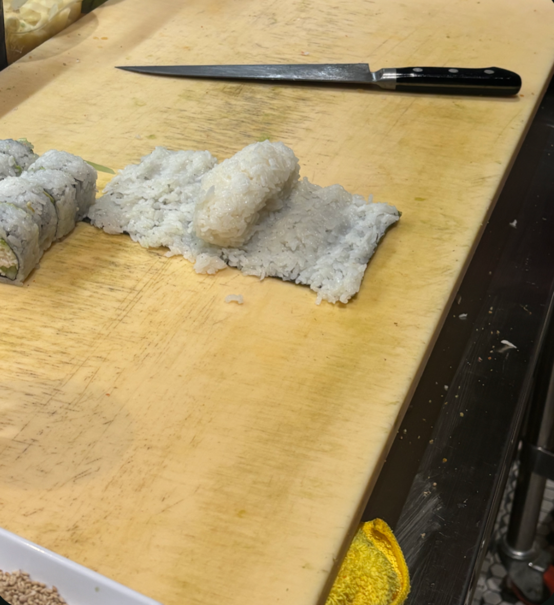 A sushi preparation area with a knife, a roll of sushi partially sliced, and pieces of white rice shaped for sushi on a wooden cutting board. A yellow cloth is visible at the edge of the board.