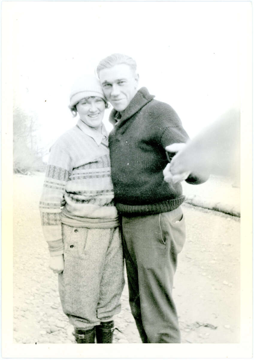 A black-and-white photo of a smiling couple standing close together outdoors; the woman wears a knit hat and sweater, and the man, in a dark sweater, has his arm around her. Another person’s hand is visible in the foreground.
