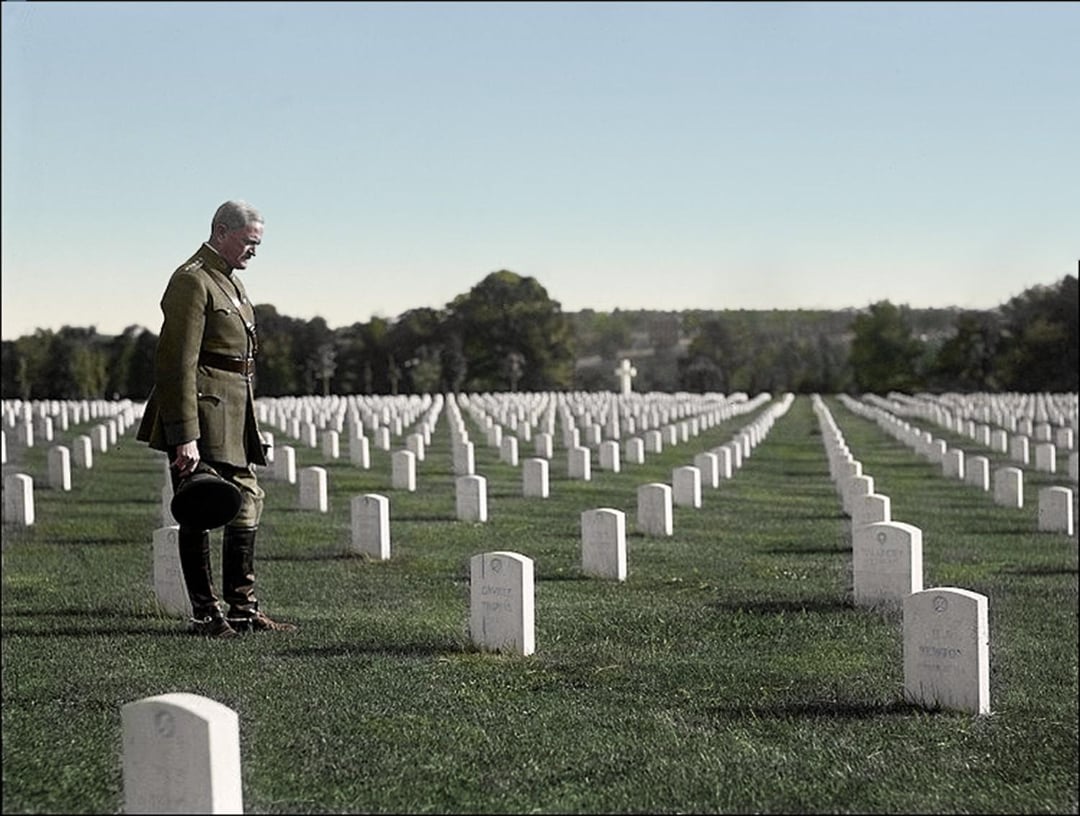 An older man in military uniform stands solemnly, holding his hat, while looking down at a headstone in a large cemetery filled with rows of white gravestones. The scene is peaceful and reflective.