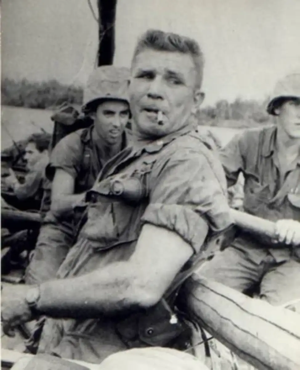 A black-and-white photo of a soldier in uniform with a cigarette in his mouth, leaning against equipment. Other soldiers in helmets are visible in the background, outdoors in a wartime setting.