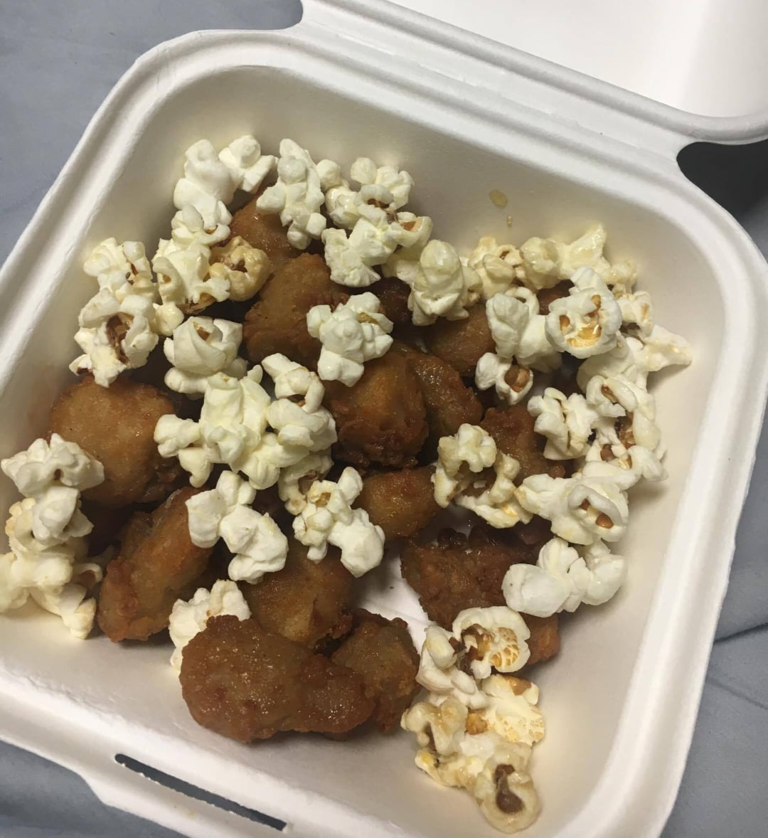 A takeout container filled with pieces of fried chicken topped with popped popcorn, placed on a light-colored surface. The lid of the container is open.
