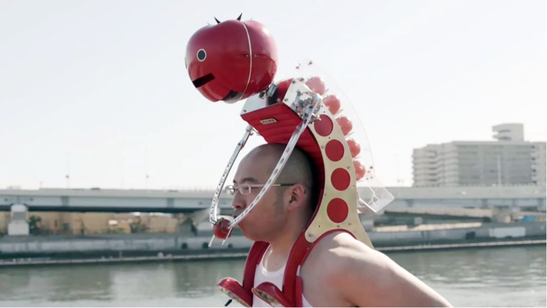 A person wearing a unique contraption with a large red tomato-shaped helmet and mechanical parts on their back stands near a body of water, with buildings and a bridge in the background.