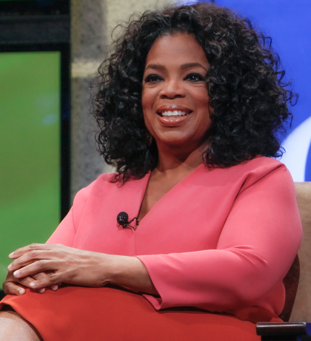 A woman with curly black hair wearing a pink and red outfit sits and smiles during a televised interview, with her hands folded on her lap.