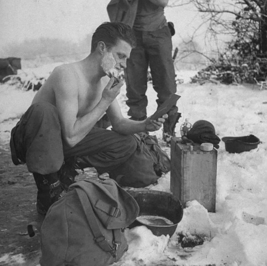 A shirtless soldier squats in the snow, shaving his face using a small mirror. Military gear and bags are scattered around, and another soldier stands in the background. The scene appears cold and wintry.