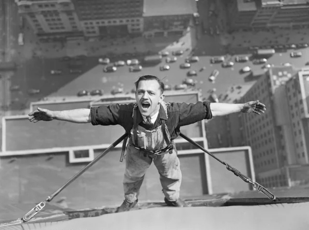A man wearing work overalls and a safety harness stands on the edge of a tall building with arms outstretched, smiling, high above a busy city street filled with cars far below.