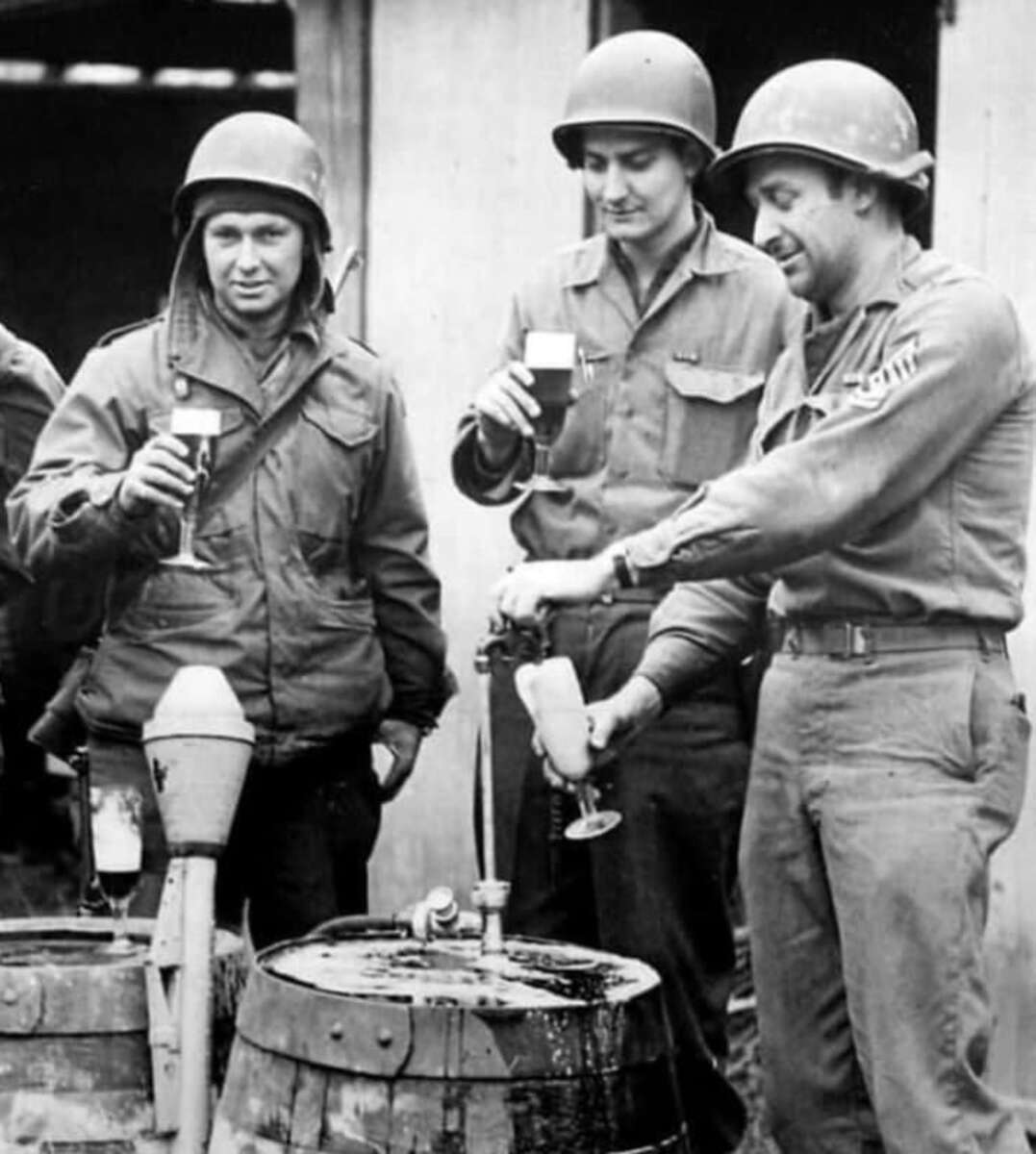 Three soldiers in World War II uniforms smile and pour beer from a barrel into glasses, standing outdoors near a wooden building. Two hold filled glasses while the third pours himself a drink.