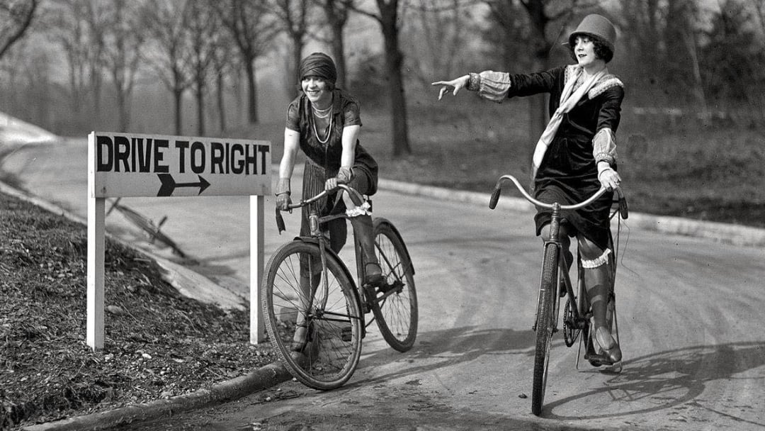 Two women in vintage clothing ride bicycles on a road next to a "DRIVE TO RIGHT" sign, with trees and a curved street in the background. One woman gestures with her arm while looking ahead.
