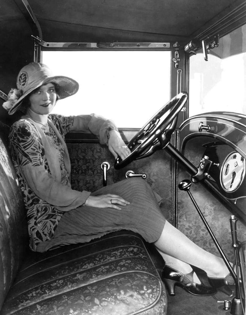 A woman in 1920s clothing and a wide-brimmed hat sits in the driver’s seat of a vintage car, smiling at the camera with one hand on the steering wheel and her legs crossed.