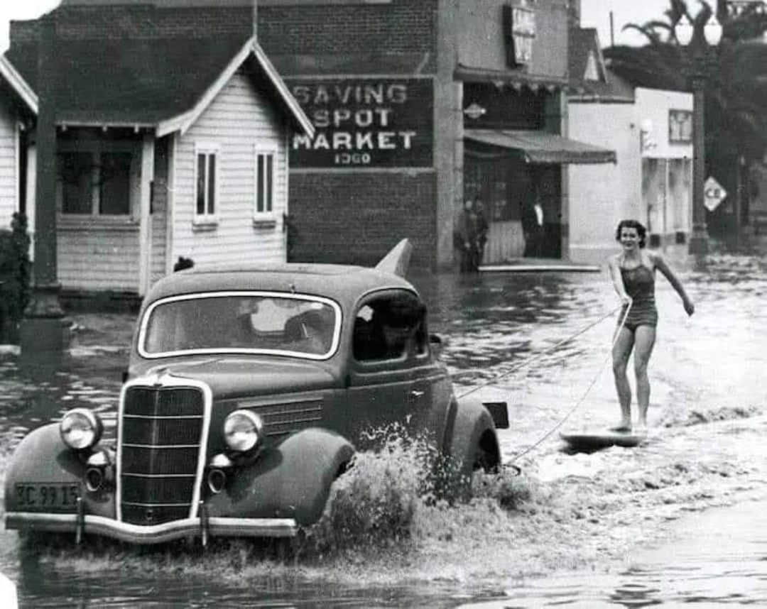 A black-and-white photo shows a woman in a swimsuit skimming over floodwaters on a board, pulled by an old car driving through a flooded street lined with houses and shops.