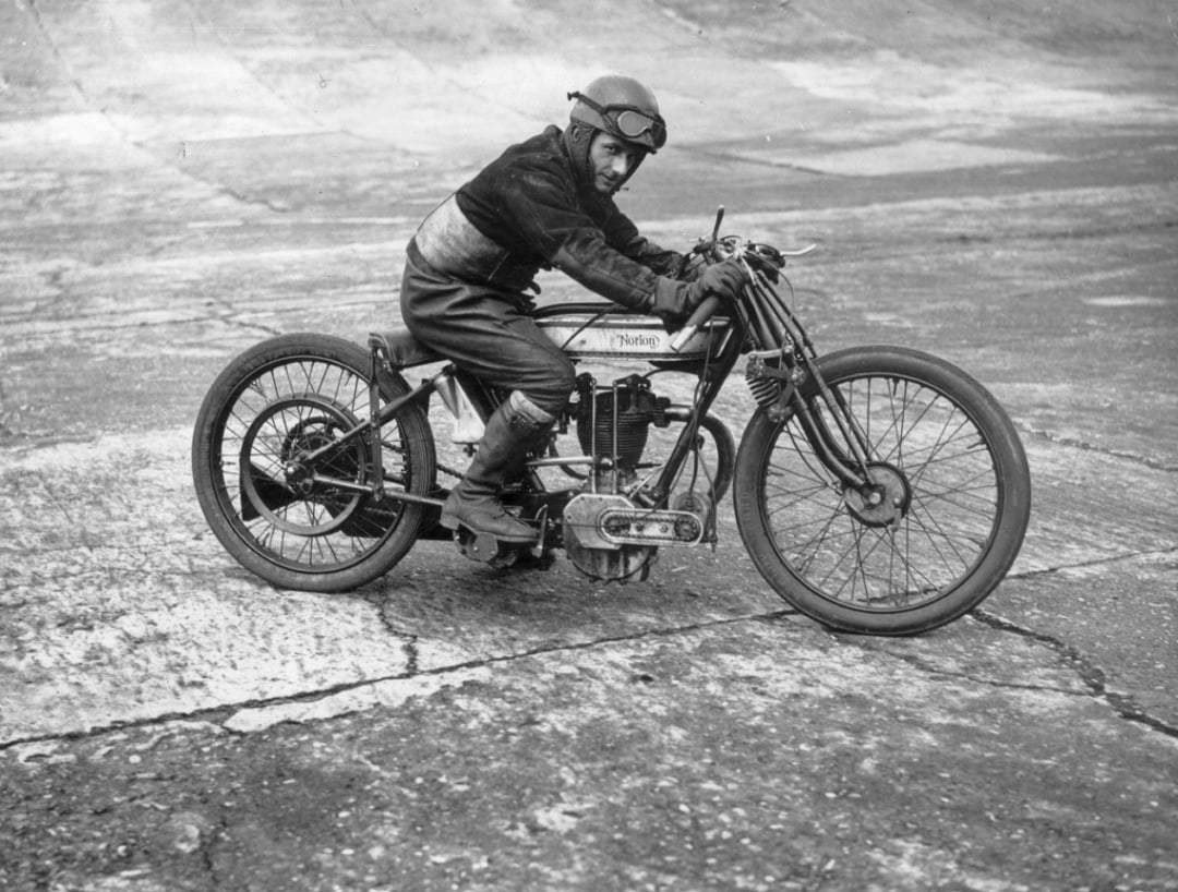 A person in vintage racing gear and goggles rides a classic Norton motorcycle on a concrete track, leaning slightly to one side while gripping the handlebars.