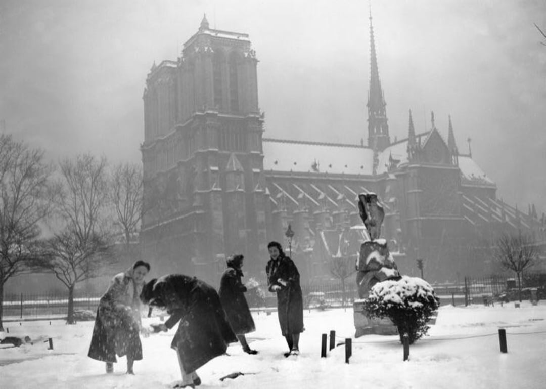 Four people play in the snow near Notre-Dame Cathedral in Paris on a snowy day. Trees and statues are dusted with snow, and the Gothic cathedral looms in the foggy background.