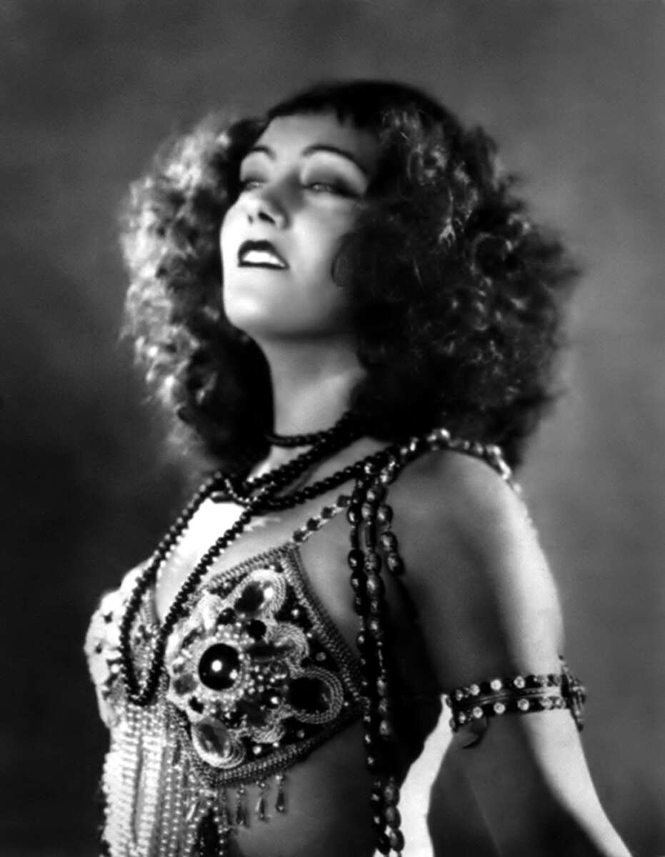 A woman with curly hair gazes upward, wearing an ornate, beaded costume with jewelry around her neck and upper arm. The photo is in black and white, giving a vintage, glamorous feel.