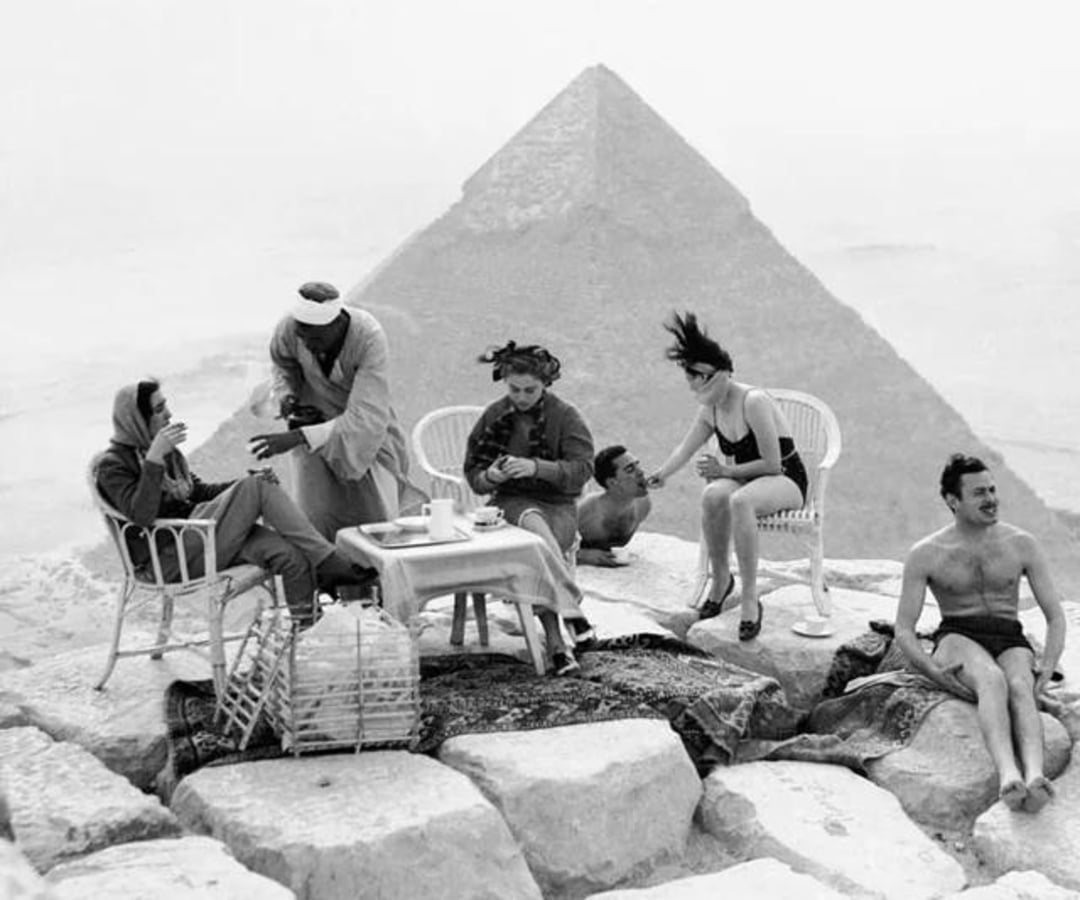 Five people relax atop stone blocks near the Great Pyramid of Giza, Egypt. Some sit in wicker chairs at a table with tea, while others wear swimsuits. A man in traditional attire serves drinks. The pyramid looms large in the background.
