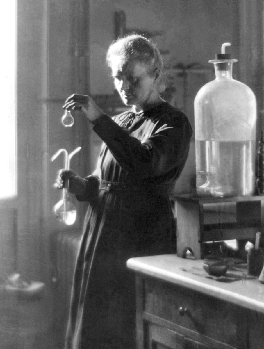 A woman in a dark dress stands in a laboratory, carefully examining a small vial she holds up in one hand while holding a flask in the other. Laboratory glassware and equipment are on the table beside her.