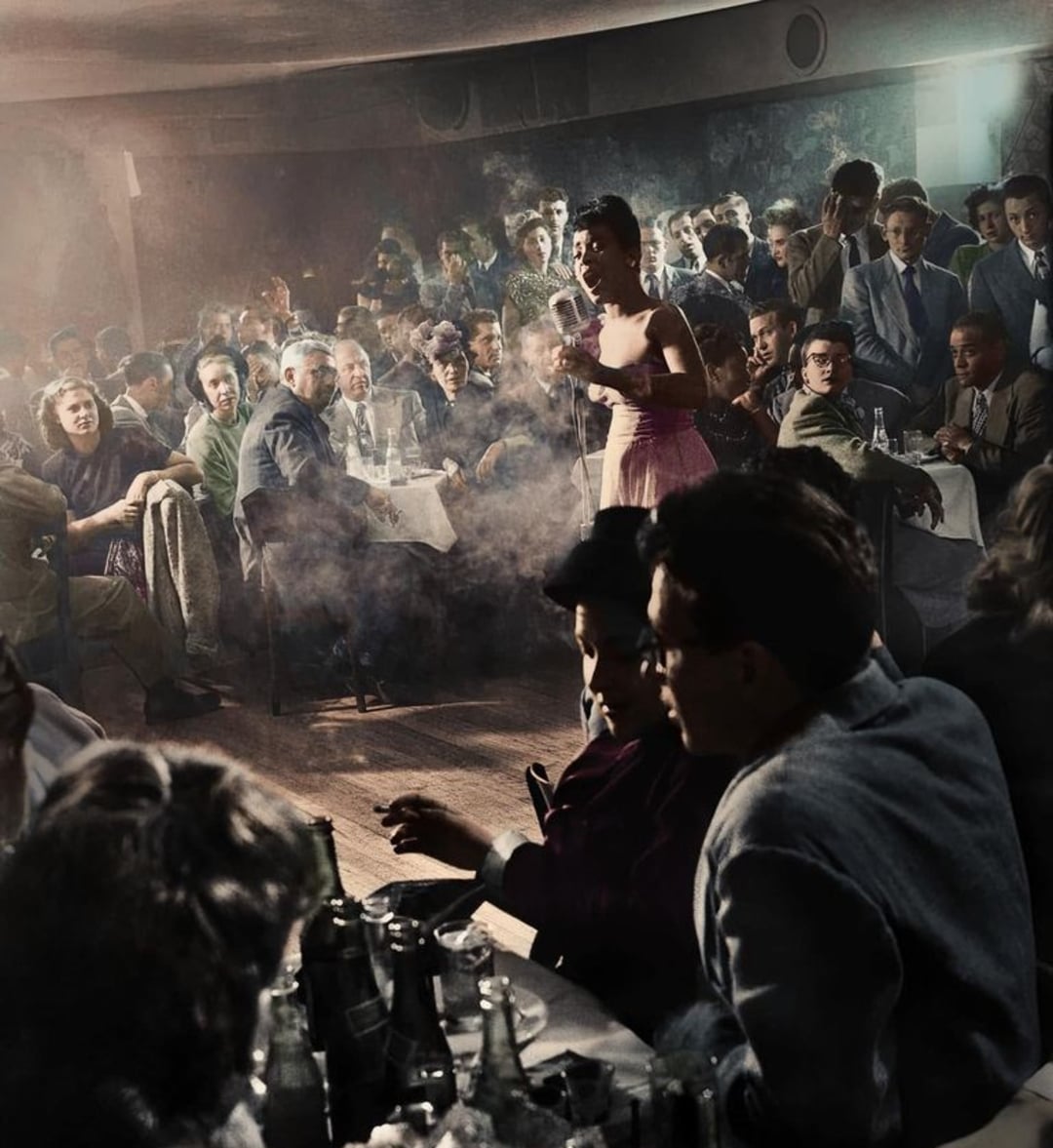 A woman in a pink dress sings on stage in a smoky, crowded jazz club, with audience members seated at tables, watching and talking among themselves. Bottles and glasses are visible on the tables in the dimly lit venue.