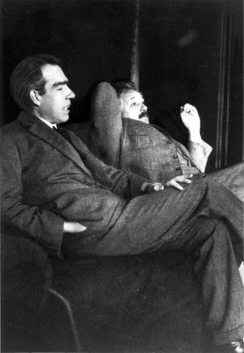 Two men in suits sitting on a couch, one with his hands in his pockets and the other leaning back with one arm behind his head, both appear to be engaged in conversation. Black and white photo.