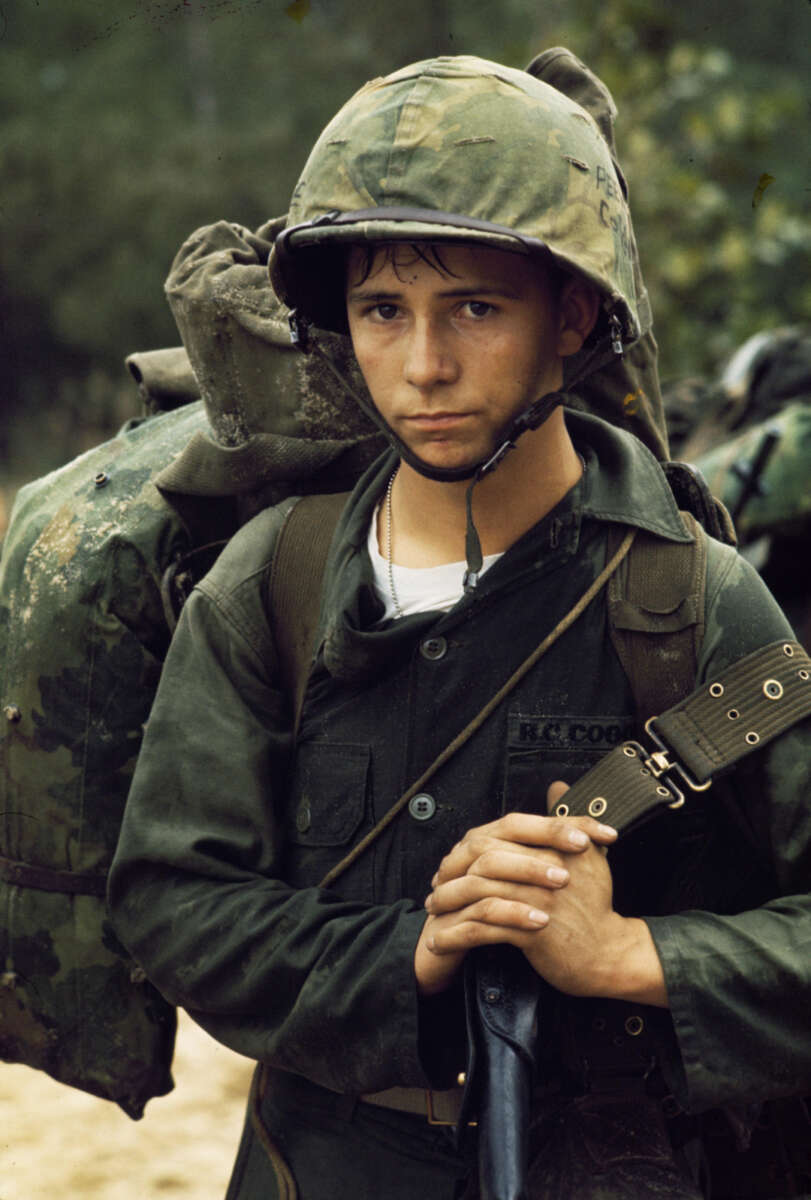 A young soldier in military uniform and helmet stands outdoors, holding a rifle. He has a serious expression and carries a large, packed backpack. The background shows greenery, suggesting a forested area.