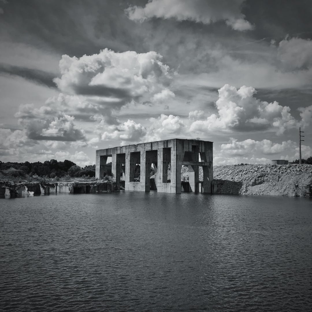 A large, abandoned concrete structure with multiple arches stands partially submerged in water under a dramatic sky filled with clouds. Rocky terrain and sparse vegetation surround the scene.