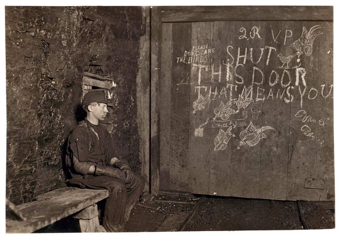 A young miner sits on a wooden bench in a dim, underground tunnel beside a large door covered with chalk drawings and warnings, including “SHUT THIS DOOR THAT MEANS YOU!”