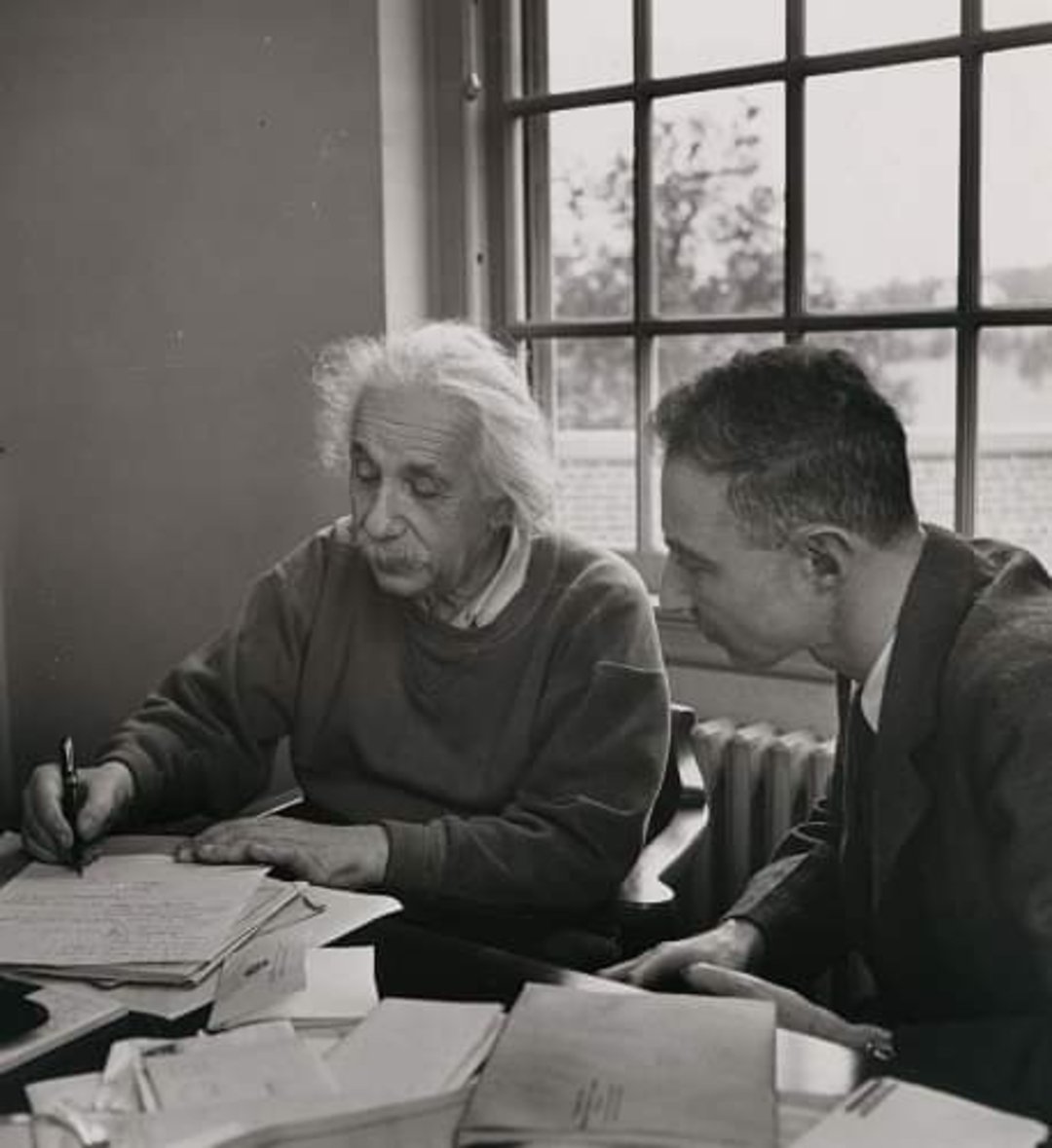 Two men sit at a cluttered desk by a window; one, with wild white hair, writes on a paper while the other leans in, watching attentively. Sunlight streams through the window behind them.