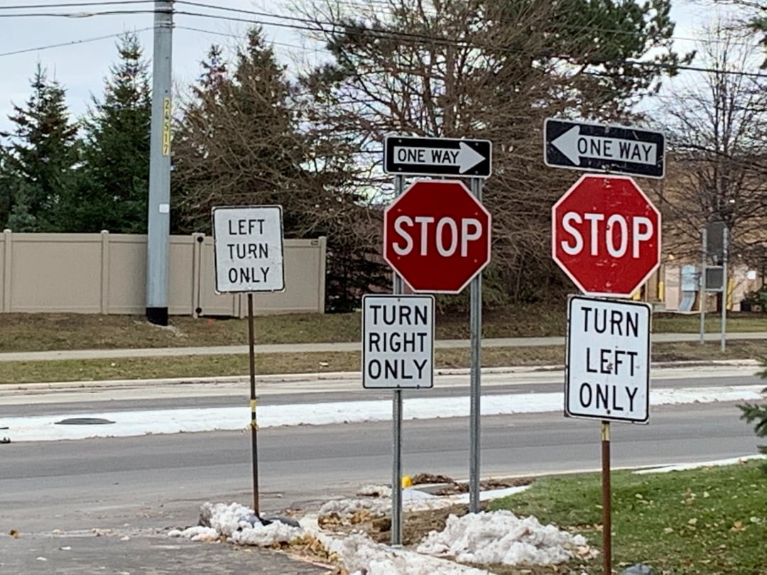 A confusing intersection with two stop signs, two "one way" signs, and conflicting "turn right only" and "turn left only" signs at the same crossing, surrounded by some snow and trees.