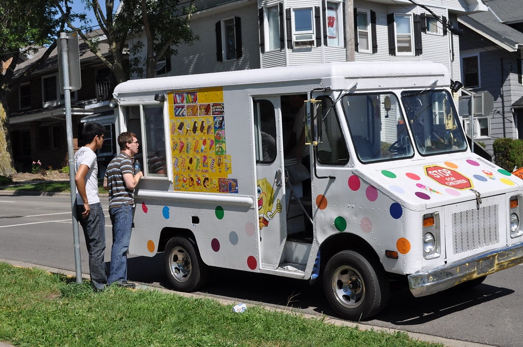 Two people stand beside a white ice cream truck with colorful polka dots, looking at its menu. The truck is parked on a residential street with houses visible in the background.
