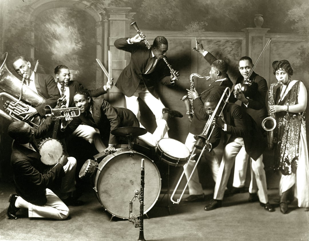 A lively jazz band poses with their instruments, including a trumpet, tuba, saxophone, drums, and clarinet. The musicians are animated, playing enthusiastically, while a woman in a flapper dress stands at the right.