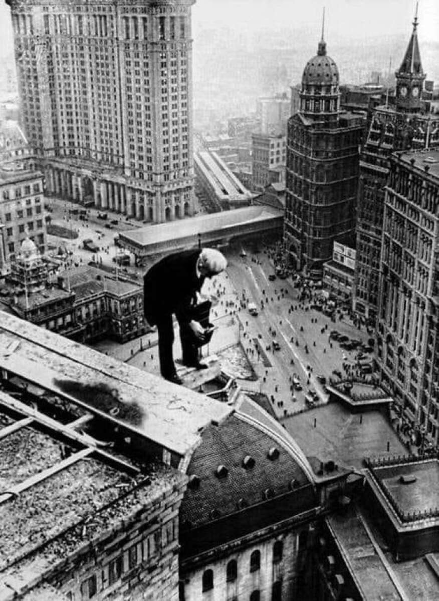 A man in a suit kneels on the edge of a tall building, looking down at a busy city street far below with people and cars, surrounded by skyscrapers and classic early 20th-century architecture.