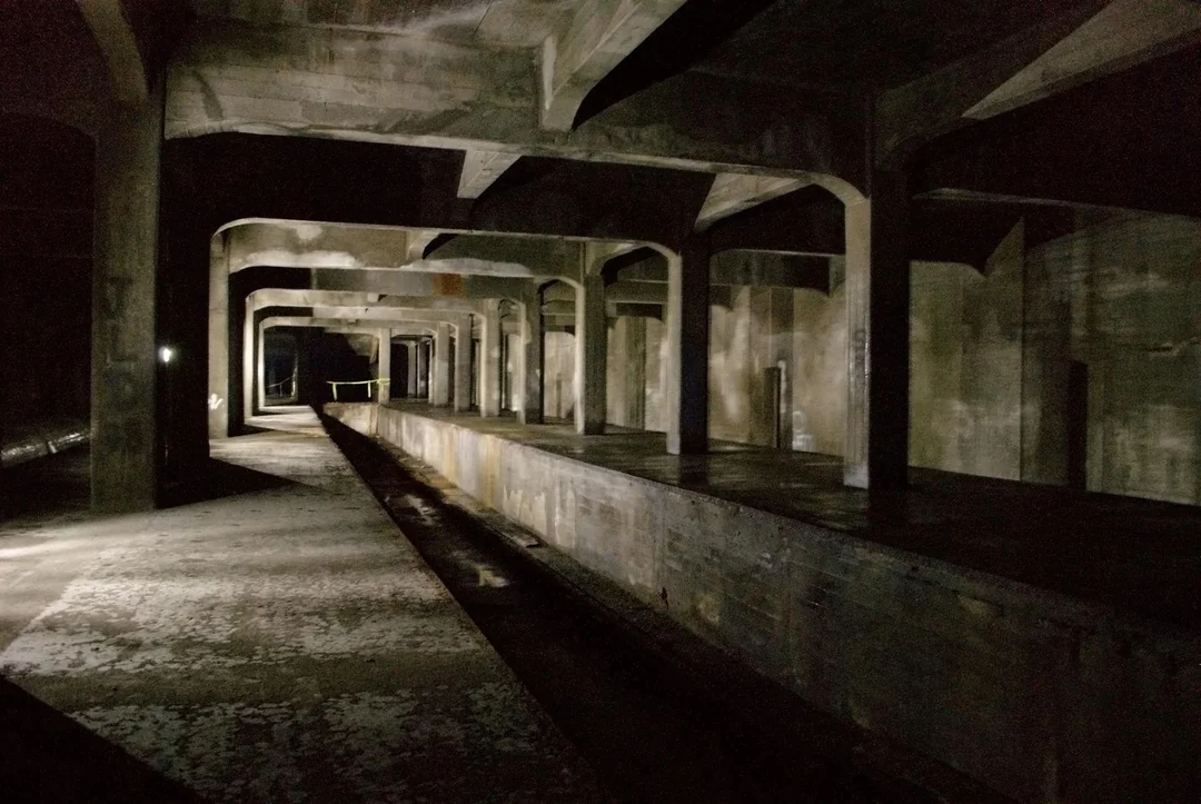 Dimly lit, abandoned concrete train platform with multiple pillars, exposed beams, and shadows. The area appears empty and weathered, creating an eerie and desolate atmosphere.