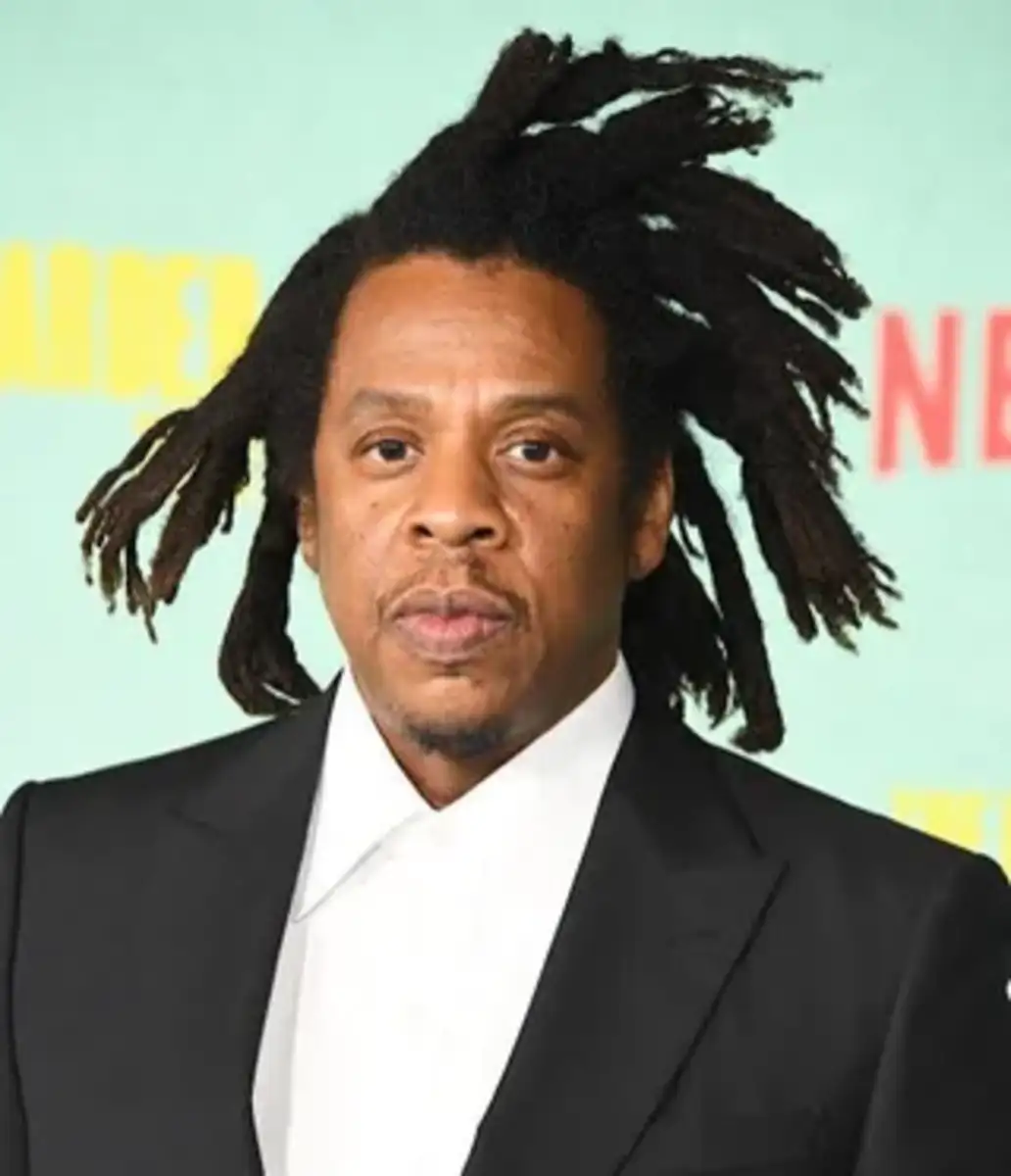 A man wearing a black suit and white shirt stands in front of a turquoise background with yellow and red text. He has medium brown skin and thick, freeform dreadlocks.