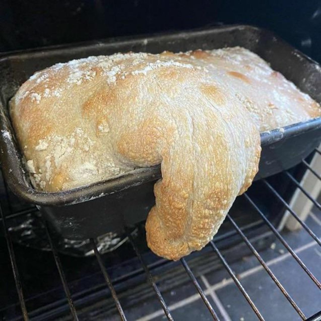 A loaf of bread has risen and overflowed from its pan while baking in the oven, with a portion of dough spilling over the edge.
