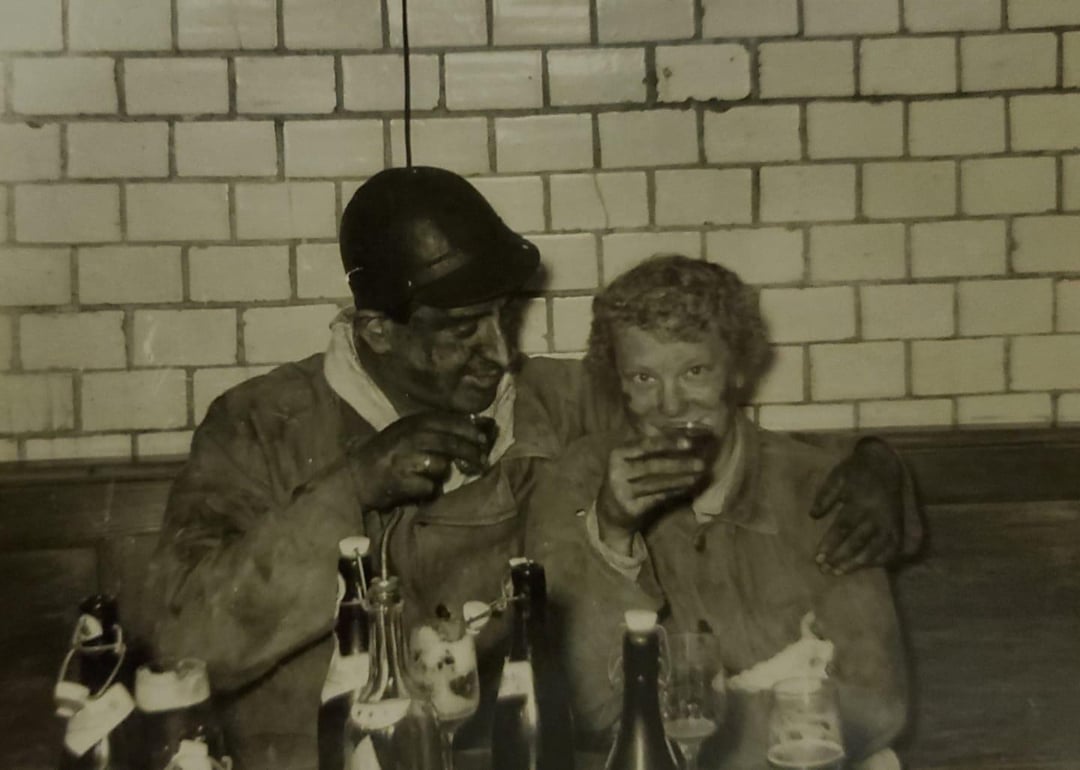 Two people in work clothes and dirty faces sit close together, smiling and holding drinks. Several bottles are on the table in front of them. They are sitting against a tiled wall, appearing cheerful and relaxed.
