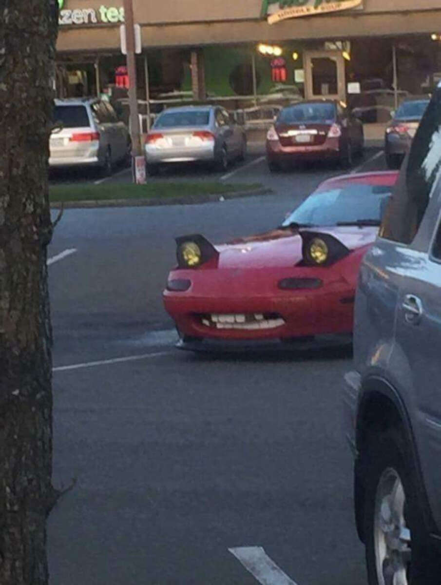 A red car is parked with its headlights up, resembling cartoonish eyes, and a fake mouth with large teeth attached to the front bumper, giving it a funny face-like appearance.