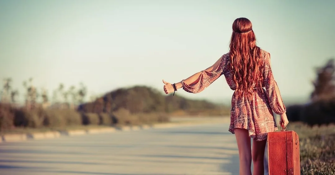 A woman with long hair wearing a patterned dress stands on the side of an empty road, holding a suitcase and extending her thumb to hitchhike under a clear sky.