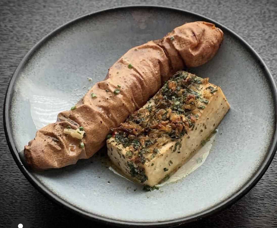 A round gray plate with a roasted sweet potato and a rectangular slice of herbed tofu garnished with chopped herbs, set on a dark surface.