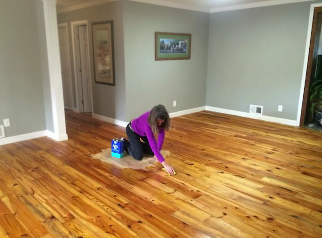 A person in a purple shirt kneels on a wooden floor, cleaning or refinishing a section of it. The room is empty, with light gray walls, white trim, and framed artwork on the walls.