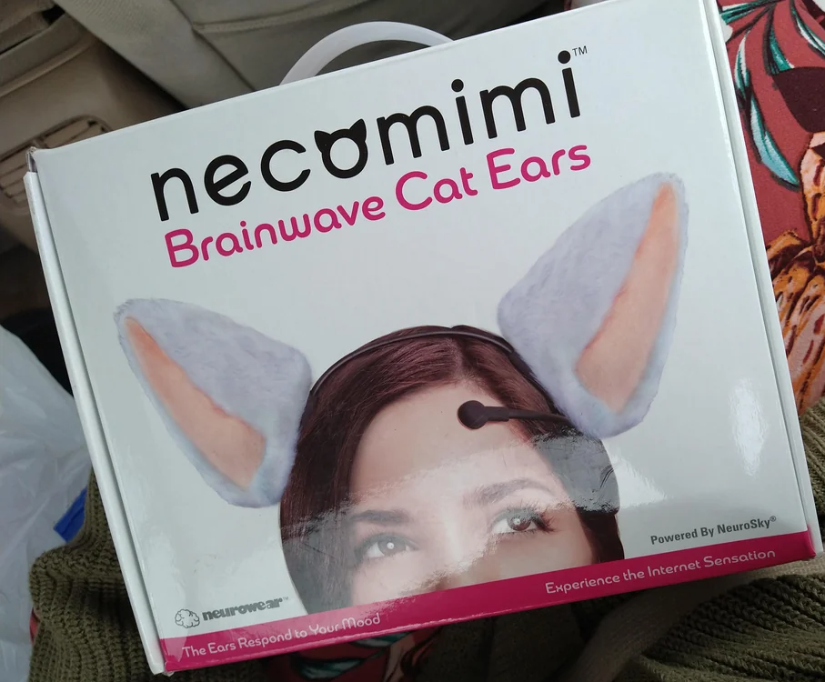 A box of "Necomimi Brainwave Cat Ears" shows a photo of a person wearing gray and pink cat ears with a sensor on their forehead. The packaging advertises that the ears respond to your mood.