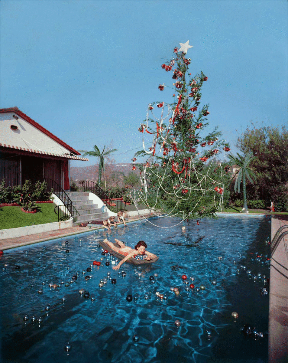 A woman lounges on an inflatable float in a swimming pool decorated with ornaments and a tall Christmas tree. The pool is outside, beside a house with a red roof, and the sky is clear and blue.