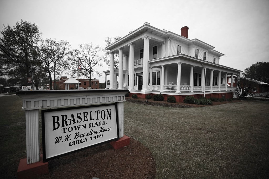 A large white historic house with columns serves as Braselton Town Hall, as indicated by a sign in front reading "Braselton Town Hall, W.H. Braselton House, circa 1909." Trees and lawn surround the building.