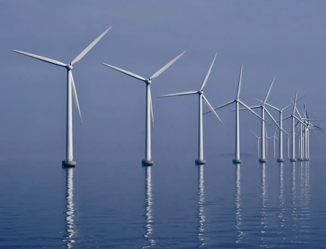 A row of large wind turbines stands in the ocean, generating renewable energy with their blades spinning above calm blue water under a hazy sky.