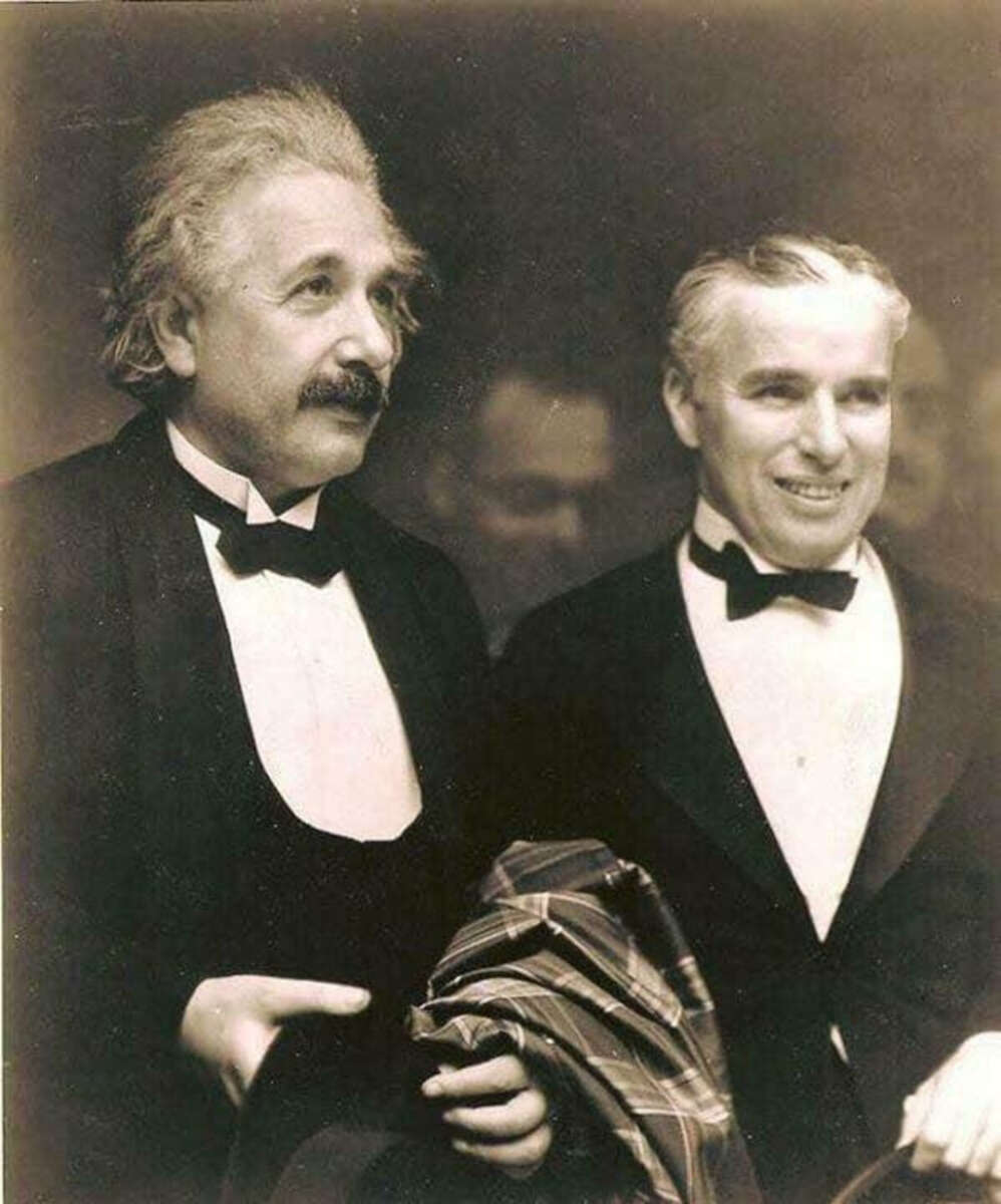 Two men in tuxedos stand side by side, smiling. One has wild, white hair and a mustache; the other has slicked, dark hair and holds a folded coat or scarf. Both appear to be at a formal event.