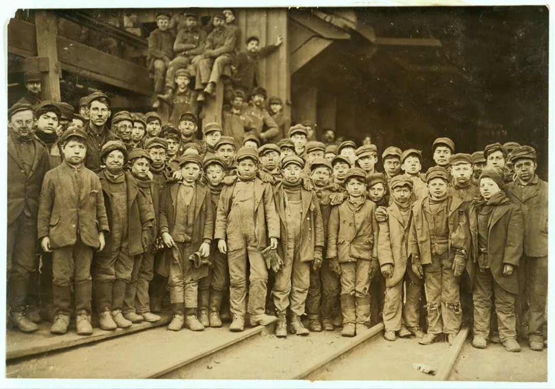 A large group of boys and men, wearing work clothes and caps, stand and sit together in front of an industrial building, possibly a coal mine, in an old sepia-toned photograph.